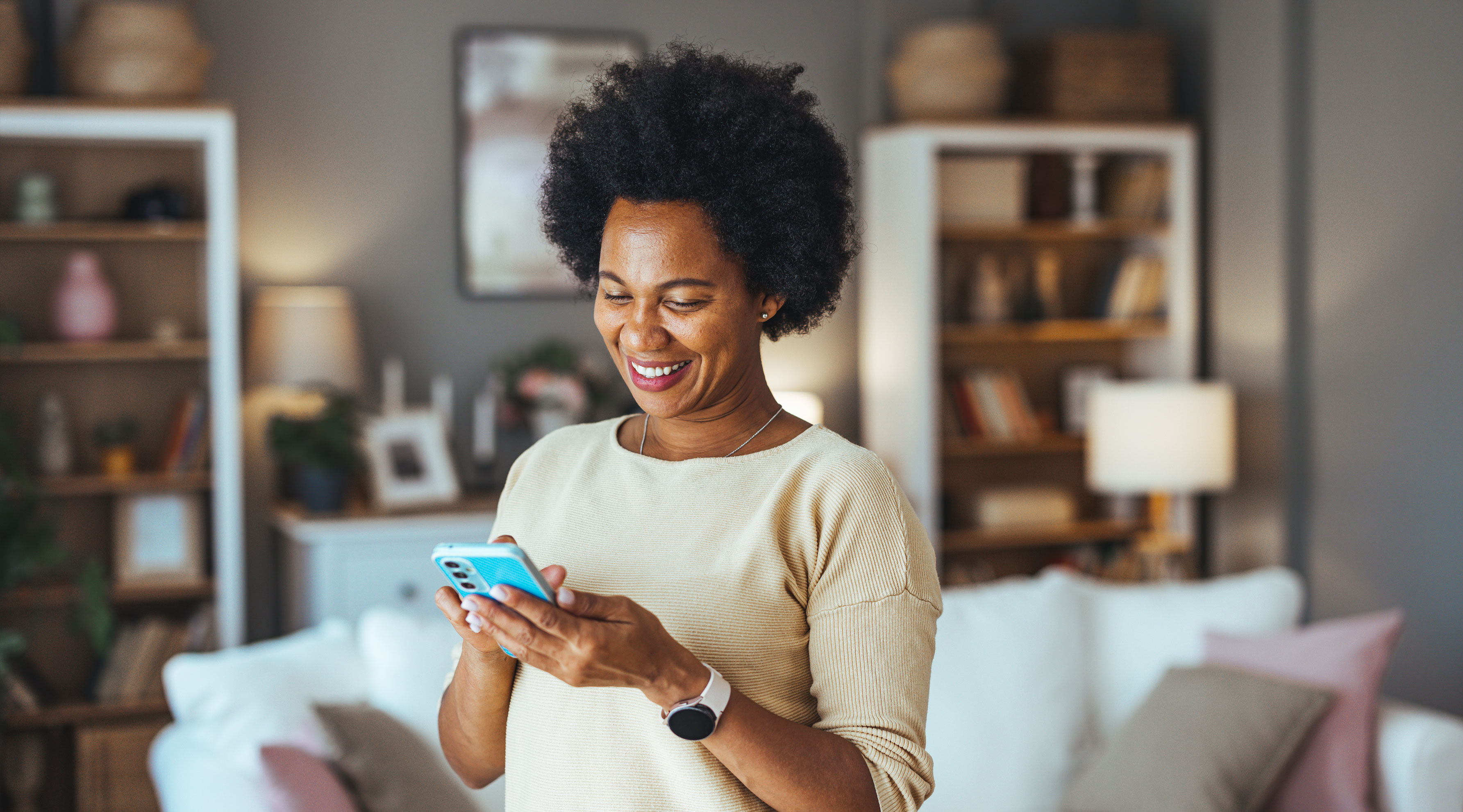 Woman with natural afro hair smiling while using her smartphone at home, wearing a cream-colored sweater and smartwatch in a cozy living room with bookshelves in the background