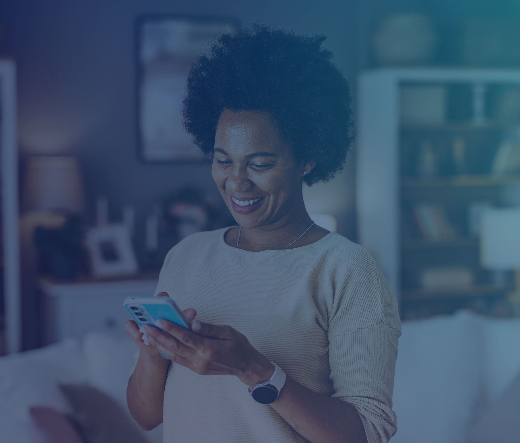 Woman with natural afro hair smiling while using her smartphone at home, wearing a cream-colored sweater and smartwatch in a cozy living room with bookshelves in the background