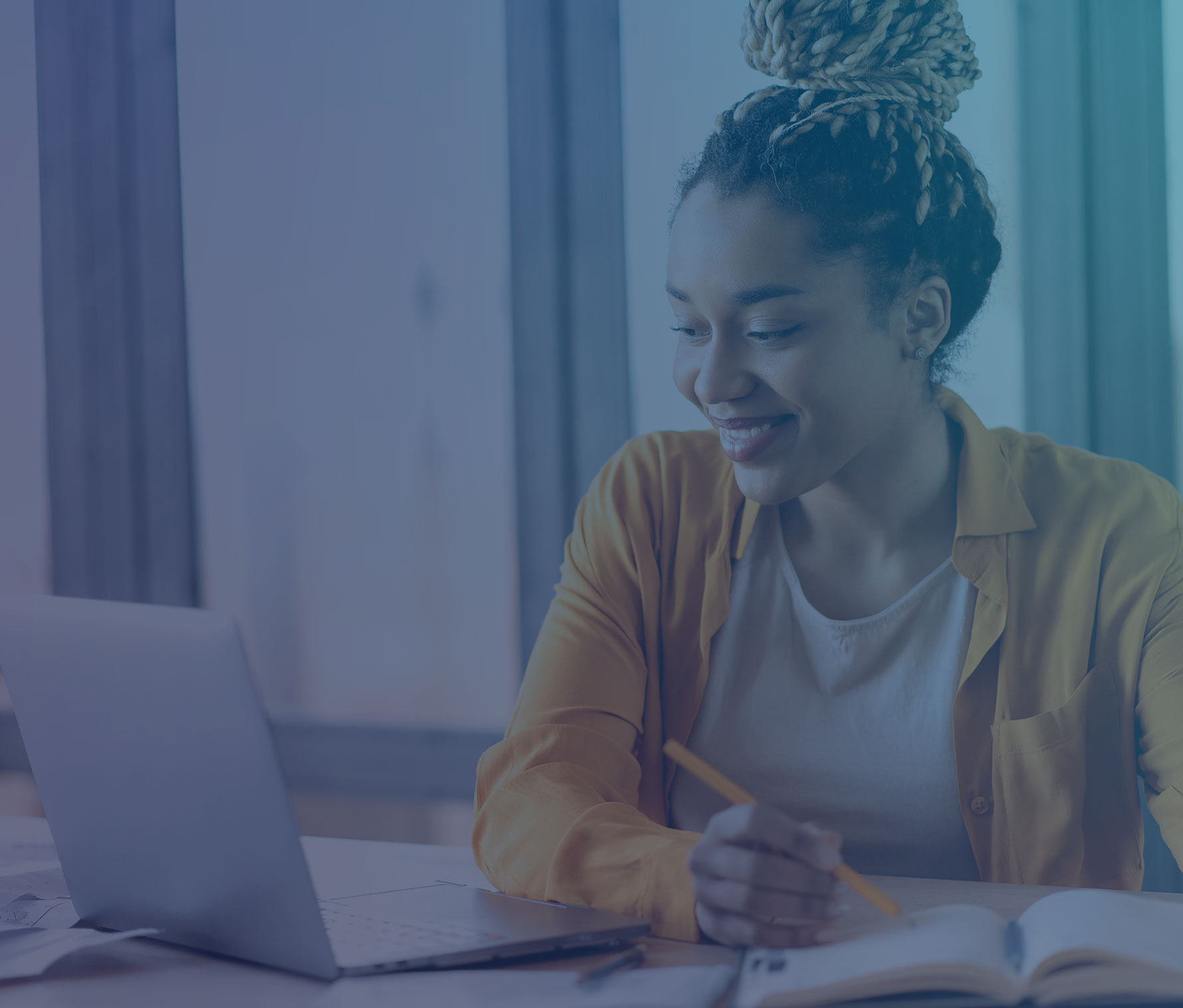 A young woman in a yellow blazer and white shirt sits at a desk, smiling while working. She has her hair styled in braids worn in a bun. In front of her is an open laptop and notebook, and she holds a pencil while studying or taking notes in a bright, window-lit space.
