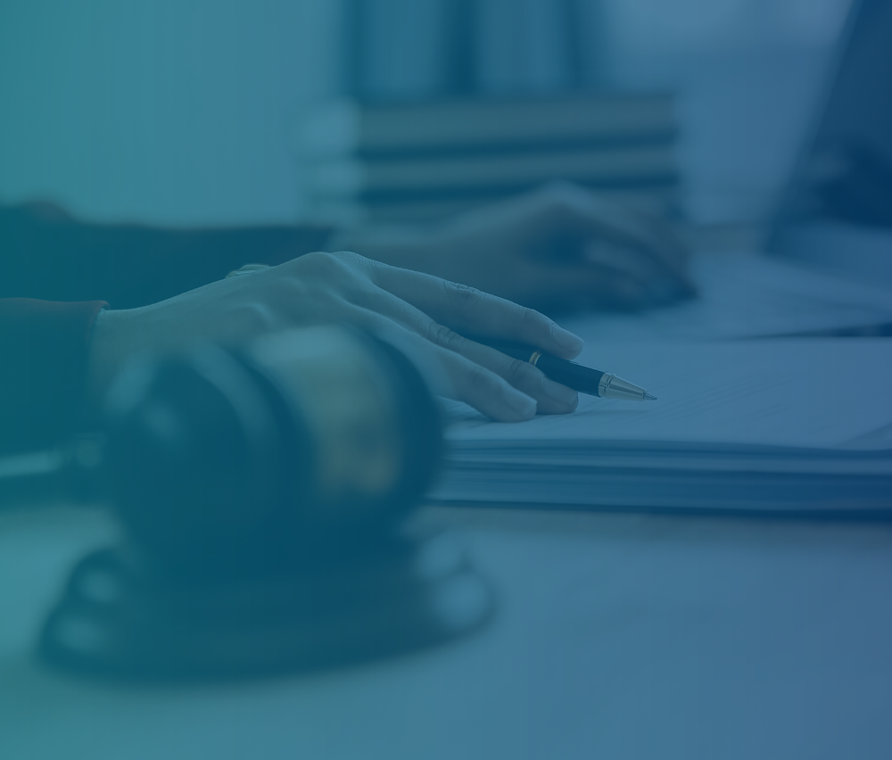 A person's hands writing on documents with a pen while using a laptop, with a wooden gavel visible on the desk, suggesting a legal work environment.